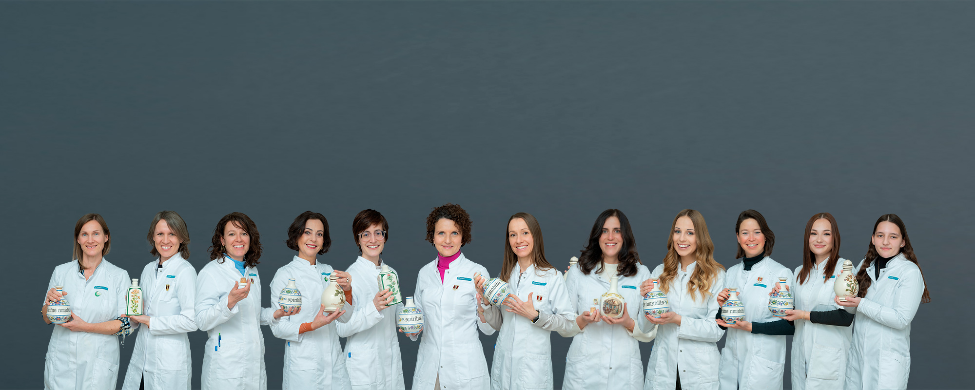 Teamfoto von 13 Apothekenmitarbeiterinnen im weißen Kittel mit verschränkten Armen vor grauem Hintergrund – geschlossen und professionell.